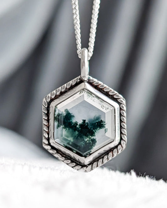 A silver hexagonal pendant necklace featuring a green forest scene gemstone, resting on a white textured surface with a gray draped fabric background.
