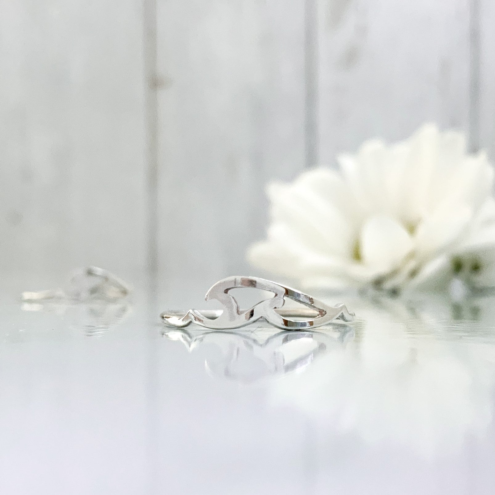 A close up of an elegant silver wave ring on a reflective surface, with out of focus white flowers behind.